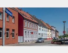 Reihe von zweistöckigen Wohnhäusern in einer Straße mit verschiedenen Fassadenfarben, darunter Rot, Grau, Blau und Beige, alle mit roten Ziegeldächern. Im Vordergrund sind Verkehrsschilder, parkende Autos und eine Blumenkiste zu sehen, der Himmel ist klar und blau.