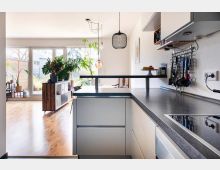 Moderne, helle Küche mit dunkler Arbeitsplatte, Einbaugeräten und Küchenutensilien an einer Wandaufhängung, die in einen offenen Wohnbereich mit Holzboden und großen, bodentiefen Fenstern übergeht; zahlreiche Topfpflanzen sind im Raum verteilt.