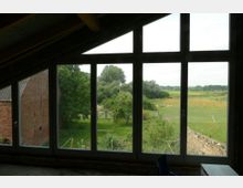 Blick durch große Fenster einer Dachgeschosswohnung auf eine ländliche Umgebung mit Wiesen, Bäumen und einem Backsteingebäude links im Bild. Im Vordergrund sind ein Teil des Innenraums und ein Tisch sichtbar.