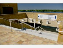 Treppenaufgang mit Geländern führt zu einem Sandstrand mit Strandkörben. Im Hintergrund befinden sich ein See, bewaldete Uferbereiche und ein blauer Himmel. Ein Schild weist zu verschiedenen Einrichtungen wie Duschen, Toiletten und Strandkorbvermietung.