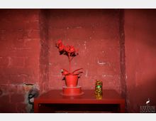 Rote Backsteinwand in einer Ecke mit roten Accessoires: ein roter Tisch, darauf ein roter Kunstblumentopf und eine goldene Winkekatze.