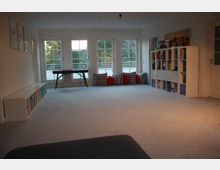 Helles Wohnzimmer mit Teppichboden, großen Fenstern und viel Tageslicht. An der rechten Wand steht ein weißes Regal mit Fächern, links ein niedriges Regal, und an den Fenstern befinden sich bunte Kissen und ein kleiner Tisch.