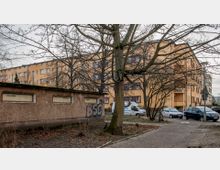 Ein Wohnhof mit einem gelb verputzten Mehrfamilienhaus im Hintergrund, davor mehrere kahle Bäume. Im Vordergrund ein flaches Gebäude mit kleinen vergitterten Fenstern und Graffiti, daneben geparkte Autos auf einem gepflasterten Weg.