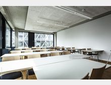 Ein moderner Klassenraum mit weißen Tischen und Holzstühlen, großen Fenstern mit Blick auf eine Gebäudefassade mit Stahlrahmen, hellem Holzboden und einer weißen Wand auf der rechten Seite. Die Decke hat ein industrielles Design mit sichtbaren Beton- und Beleuchtungselementen.