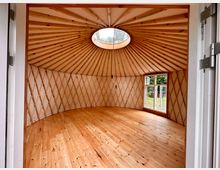 Innenraum einer runden Jurte mit Holzfußboden, hellen Wänden und einem kuppelförmigen Dach mit sichtbaren Holzstreben. Im Dach befindet sich eine runde Öffnung, und an der Seite sind Fenster mit Blick ins Grüne.