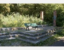 Eine grüne Parkbank steht auf einer leicht erhöhten, mit Steinplatten bedeckten Plattform, umgeben von niedrigen Stufen. Im Hintergrund befinden sich dichte Vegetation und zwei gemauerte Säulen aus roten Ziegeln.