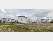 Mehrstöckige, moderne Wohngebäude mit hellen Fassaden stehen am Rand einer offenen Grünfläche. Ein Werbeschild für Neubauprojekte ist im Vordergrund sichtbar, und mehrere Bäume und parkende Autos befinden sich entlang des Weges zwischen den Gebäuden.