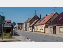 Straßenszene in einer kleinen Ortschaft mit niedrigen Wohnhäusern in verschiedenen Farbtönen, teils mit roten Ziegeldächern; einige Gebäude haben Solarpanels. Links im Bild befinden sich Wegweiser und ein Gehweg mit etwas grünem Bewuchs.