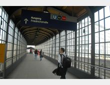 Ein überdachter Fußgängerkorridor mit Glaswänden und einem Dach aus Holz und Metall, der zu einem Ausgang mit der Beschilderung "Ausgang Friedrichstraße" führt; mehrere Personen gehen Richtung Ausgang.