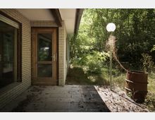 Außenansicht eines verlassenen Gebäudes mit Ziegelwänden und einer Holztür, umgeben von dichter Vegetation. Im Vordergrund sind eine verrostete Tonne und eine Laterne mit einer überwucherten Rankpflanze zu sehen.