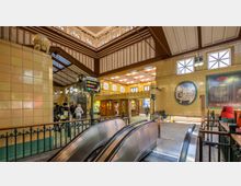 Historische U-Bahn-Station mit gelb gekachelten Wänden, verzierten Holzelementen, großen Fensterfronten und einer Rolltreppe im Vordergrund. Im Hintergrund befinden sich Wegweiser zu den Bahnsteigen sowie Personen, die durch die Station gehen.