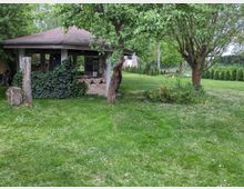 Garten mit Rasenfläche, mehreren Bäumen und einem offenen Pavillon aus Holz und Ziegeln, teilweise von Efeu bewachsen. Im Hintergrund sind Büsche und eine Hecke zu sehen, die das Grundstück begrenzen.