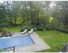 Garten mit einem rechteckigen Pool, der mit einer blauen Abdeckung bedeckt ist, umgeben von einer Holzterrasse. Im Hintergrund befinden sich eine Rasenfläche, große Bäume und eine erhöhte Holzplattform mit Geländer, die über eine Treppe erreichbar ist.