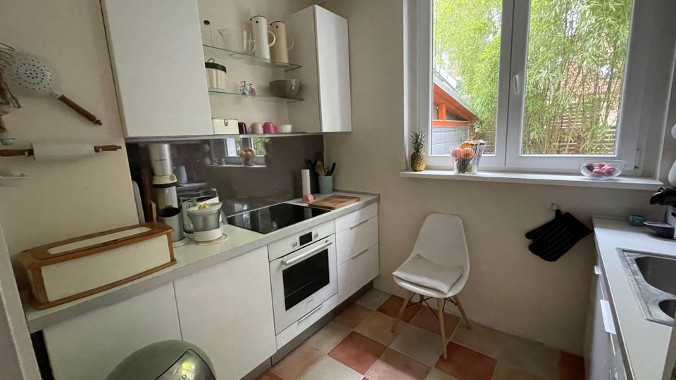 Kleine, helle Küche mit weißen Einbauschränken, Arbeitsplatte mit Herd und Backofen sowie offenem Regal und Edelstahl-Rückwand. Großes Fenster mit Blick ins Grüne, gefliester Boden in Beige- und Terrakottatönen, dazu ein Stuhl neben der Fensterbank und eine Spüle auf der rechten Seite.