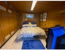 Ein kleines Schlafzimmer mit Holzvertäfelung an Wänden und Decke. Zentral steht ein Bett mit bunter Blumenbettwäsche und einem Stofftier darauf, daneben ein kleiner Nachttisch; links befinden sich Holzschränke, und im Vordergrund ist ein blauer Stuhl und ein TV-Gerät sichtbar.