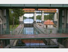 Blick auf eine historische Schleusenanlage mit mehreren Ebenen, einem zentralen Wasserbecken und grünen Metallgeländern. Im Hintergrund sind eine Wasserstraße, Bäume und ein Gebäude mit roten Ziegeldächern zu sehen.