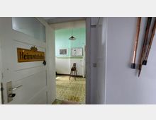 Blick durch eine geöffnete Holztür mit der Beschriftung „Heimatstube“ in einen Raum mit türkisgrünen Wänden, einer einfachen Deckenlampe und einer alten Holzbettstelle. An den Wänden hängen Bilder sowie Infotafeln, und rechts im Vordergrund sind Spazierstöcke an der Wand befestigt.