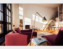 A spacious, modern living room with large floor-to-ceiling windows, burgundy seating, and a minimalist fireplace next to stacked firewood. The room features a staircase leading to a loft area with extensive bookshelves, wooden floors, and mid-century style furniture.