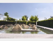 Flacher Wasserbrunnen mit modernen, terrassenförmigen Steingebilden in einem rechteckigen Becken, umgeben von Hecken und vereinzelten Bäumen, unter einem klaren blauen Himmel. Im Hintergrund ist eine niedrige Mauer zu erkennen.