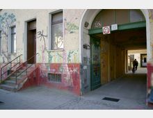 Fassade eines Gebäudes mit Wandmalereien und Graffiti, links eine kleine Treppe mit Metallgeländer, rechts ein Durchgangsbogen mit einer Fahrradfahrerin im Hintergrund. Die Hausnummern 25 und 39 befinden sich über dem Durchgang.