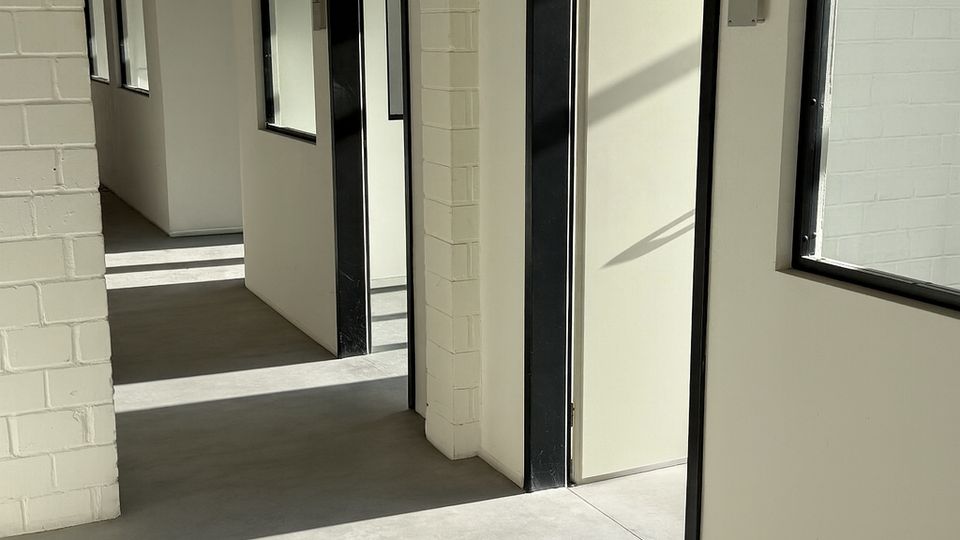 Heller Innenflur in einem modernen Gewerbe- oder Bürogebäude mit weißen Wänden und sichtbaren Ziegelpfeilern, grauem Boden sowie Türen und Fenstern mit schwarzen Rahmen. Sonnenlicht fällt durch die Fenster und wirft lange Schatten auf den Boden.