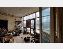 Ein einfacher Werkstattraum mit abgenutzten Wänden, Regalen mit Werkzeugen und Materialien sowie gestapelten Gegenständen. Eine große Fensterfront mit Metallgittern und einer Tür lässt Tageslicht herein, während draußen ein Hof mit einem geparkten Auto sichtbar ist.