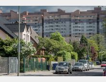 Wohnstraße mit Einfamilienhäusern, Vorgärten und geparkten Autos, im Hintergrund ein großes, mehrstöckiges Wohngebäude aus Beton mit Fensterrastern. Die Straße ist von Bäumen und Sträuchern gesäumt.
