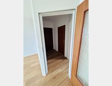 Blick durch einen Türrahmen in einen kleinen, hellen Wohnungsflur mit Holzlaminatboden und weißen Wänden. Im Flur sind zwei dunkle Holztüren sichtbar, rechts im Vordergrund eine geöffnete Tür mit strukturiertem Glaseinsatz.
