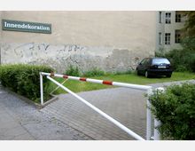 Hinterhof mit einem gepflasterten Weg, einer rot-weiß gestrichenen Schranke, angrenzenden Grünflächen und einem geparkten Auto. Im Hintergrund eine verwitterte Hauswand mit verblasstem Graffiti und einer Beschilderung „Innendekoration“.