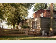 Holzhaus mit Ziegelaufbau, umgeben von Bäumen und Büschen. Im Vordergrund ein niedriger Holzzaun mit einem Tor, dahinter befindet sich ein ruhiges, ländliches Grundstück.