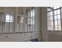 Innenraum mit weißen, holzgerahmten Sprossenfenstern, die den Raum auf zwei Seiten einrahmen. Unter den Fenstern befindet sich eine holzverkleidete Wand, ein Heizkörper und vereinzelte sichtbare Kabelanschlüsse.