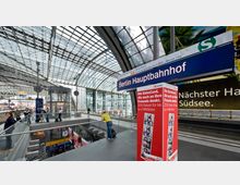 Innenansicht des Berliner Hauptbahnhofs mit moderner Glas- und Stahlarchitektur; im Vordergrund ein Hinweisschild mit der Aufschrift "Berlin Hauptbahnhof" und eine rot-weiße Werbesäule. Menschen bewegen sich entlang einer Galerie mit Blick auf Bahnsteige und mehrere Ebenen darunter.