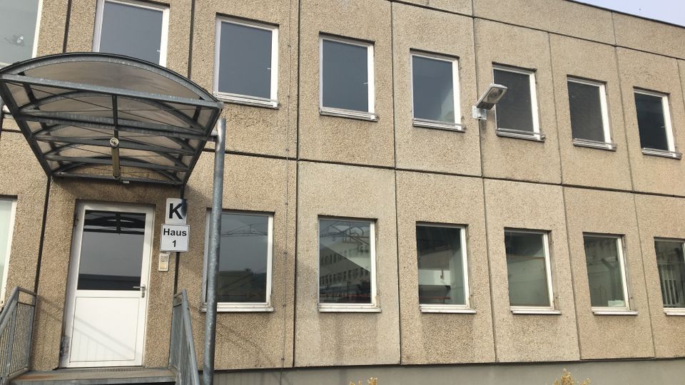 Zweistöckiges, schlichtes Büro- oder Verwaltungsgebäude mit beigefarbener Betonfassade und Reihen rechteckiger Fenster. Links ein Eingang mit Treppe und gebogenem Glasvordach; daneben ein Schild „K Haus 1“ und eine Außenleuchte an der Fassade.