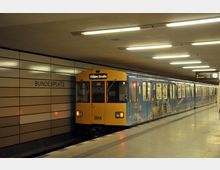 U-Bahn-Station mit gelb-blauer U-Bahn, die mit der Zielanzeige „Osloer Straße“ am Bahnsteig hält. An der Wand steht „Bundesplatz“, und die Beleuchtung ist hell und gleichmäßig.