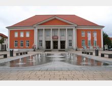 Kulturzentrum mit orangeroter Fassade und weißen Säulen im Eingangsbereich, davor eine gepflasterte Fläche mit kleinen, sprudelnden Wasserspielen. Links und rechts sind Sitzbänke und Informationstafeln aufgestellt.