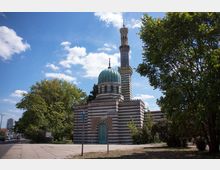 Historisches Gebäude mit Kuppel und Minarett in Streifenmuster-Architektur, umgeben von Bäumen und einem Gehweg, unter einem sonnigen Himmel mit vereinzelten Wolken. Im Hintergrund sind eine Straße mit Autos und moderne Gebäude zu sehen.
