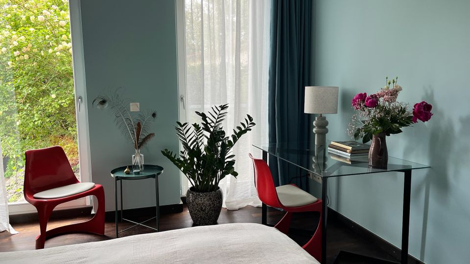 Modernes Schlafzimmer mit hellblauen Wänden, bodentiefen Fenstern mit transparenten Vorhängen und Blick ins Grüne. Im Raum stehen ein Glas-Schreibtisch mit Lampe und Blumen, rote Stühle, ein kleiner Beistelltisch und mehrere Zimmerpflanzen auf dunklem Holzboden.