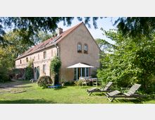 Zweistöckiges Backsteingebäude mit roten Dachziegeln und Doppelfenstern, umgeben von Bäumen und Grünflächen. Im Vordergrund befinden sich Liegestühle, ein Tisch mit Sonnenschirm sowie ein gepflegter Garten.