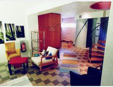 Wohnraum mit gemütlicher Einrichtung, darunter ein gelber Sessel, ein Schaukelstuhl und ein kleiner Tisch mit roter Tischdecke auf einem gefliesten Boden. Im Hintergrund befinden sich eine Holzverkleidung, ein Wendeltreppenaufgang aus Holz und eine ovale gemusterte Duschkabine; bunte Teppiche liegen verstreut auf dem Boden.