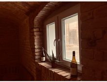 Fenster in einem Raum mit Ziegelwänden und gewölbter Ziegeldecke, mit zwei Weinflaschen und einer kleinen Pflanze auf der Fensterbank. Durch das Fenster ist ein Außenbereich mit weiteren Ziegelmauern sichtbar.