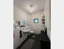 Modernes Badezimmer mit weißen Wandfliesen, einem kleinen Fenster mit mattiertem Glas, einem Waschbecken und einer Hänge-Toilette. Der Raum ist mit Spiegeln, Regalen und schwarzen Möbeln dekoriert, der Boden ist dunkel gefliest.
