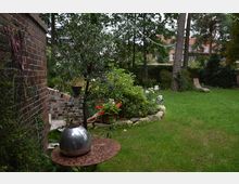 Grüner Garten mit Rasenfläche, Blumenbeeten und Bäumen, daneben ein gemauerter Backsteinbau. Im Vordergrund steht ein metallischer Kugel-Dekoartikel auf einem verrosteten Tisch, im Hintergrund ist eine braune Liege und ein Wohngebäude mit roten Dachziegeln zu sehen.