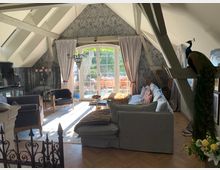 Wohnzimmer in einem Dachgeschoss mit sichtbaren Holzbalken, einer großen Fensterfront mit Blick auf eine Terrasse, grauer Tapete mit Muster und hellgrauen Vorhängen. Der Raum ist eingerichtet mit einem grauen Sofa, Sesseln, einem Couchtisch und dekorativen Elementen wie einem Kronleuchter, Kerzenständer und einem ausgestopften Pfau.