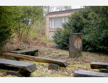 Eine kleine, von Bäumen und Sträuchern umgebene Freifläche mit Holzbänken und einem Baumstumpf, in den ein Kreuzsymbol geschnitzt ist; im Hintergrund ist ein einfaches Backsteingebäude mit vergitterten Fenstern zu sehen.