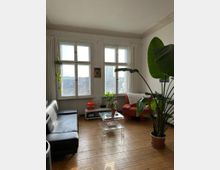 Helles Wohnzimmer mit Holzboden, großen Fenstern und weißem Vorhang. Möbel umfassen ein schwarzes Sofa, einen roten Sessel, einen transparenten Couchtisch und Zimmerpflanzen, darunter eine große Grünpflanze im Vordergrund.