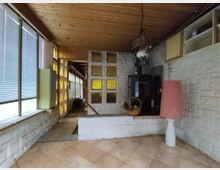 Ein rustikaler Wohnraum mit heller Kachelfußboden, Holzdecke und weiß gestrichenen Mauerwänden. Im Raum befinden sich ein Kaminofen, eine Hängelampe, ein Korb mit Holzscheiten und eine große Stehlampe mit rotem Schirm; gelbe Glasbausteinwände trennen Bereiche ab, und bodentiefe Fenster mit Jalousien lassen Tageslicht hinein.