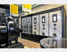 Ein historischer Maschinenraum mit großen schwarzen Industrieanlagen im Vordergrund und einer Wand aus Schalt- und Messgeräten im Hintergrund. Der Raum hat einen rot-weißen Fliesenboden und ist mit gelben Schildern über technisches Kulturerbe ausgestattet.