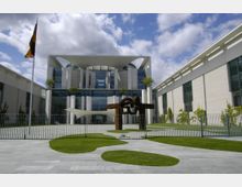 Das Bild zeigt das Bundeskanzleramt in Berlin, ein modernes Regierungsgebäude mit einer symmetrischen Fassade und markanten Beton- und Glasarchitekturelementen. Im Vordergrund befindet sich eine große Skulptur aus Metall, umgeben von einem gepflegten Garten mit Rasenflächen und gepflastertem Weg.