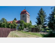 Dorfkirche mit einem steinernen Turm und rotem Ziegeldach, umgeben von Bäumen und Gärten; im Vordergrund ein unbefestigter Weg und ein Holzzaun. Der Himmel ist klar und blau.