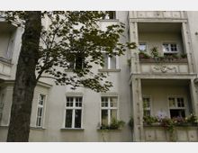 Mehrstöckiges Wohngebäude mit hellen Fassaden und verzierten Balkonen. Vor dem Gebäude steht ein Baum mit grünen Blättern, und einige Fenster und Balkone sind mit Pflanzen geschmückt.